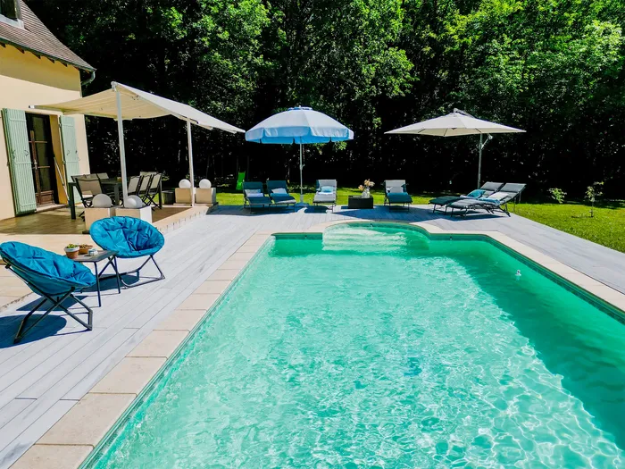 Terrasse en bois composite autour de la piscine, fauteuils bleus et volets verts de la maison.