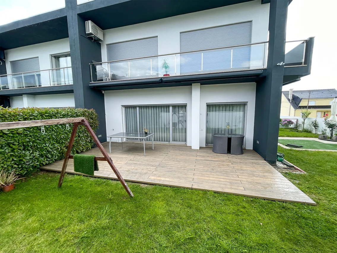 Terrasse composite Natural Beech vue depuis le jardin, balancelle en bois, table et fauteuils en résine tressée devant la façade à balcon.