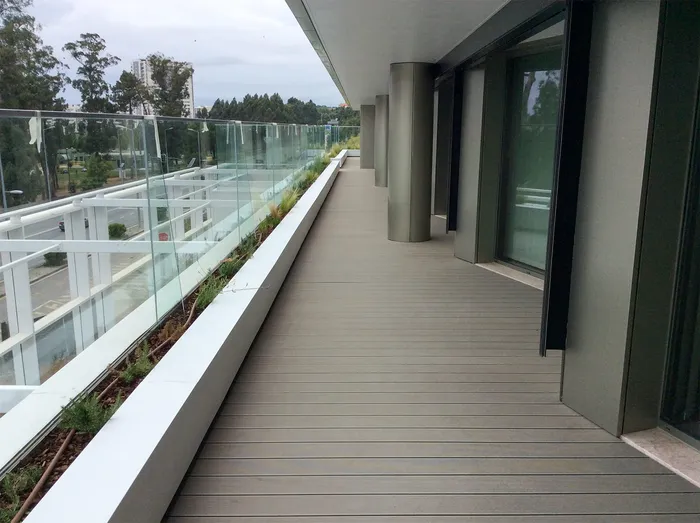 Long balcony in grey composite decking along the façade, with metal pillars, a planter and a glass balustrade.