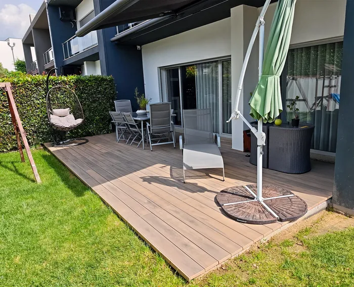 Terrasse composite CDECK® Natural Beech agrandie en jardin arrière, fauteuil suspendu, salon repas et bain de soleil sous un parasol.
