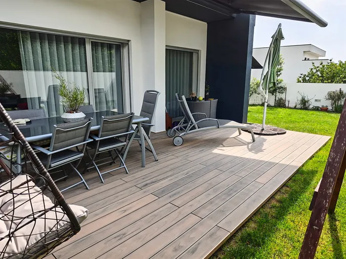 Vue d'ensemble de la terrasse composite Natural Beech bordée de pelouse, salon de jardin et parasol replié.