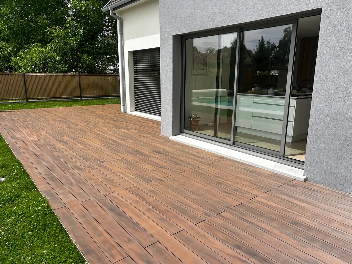 Terrasse composite CDECK WUUDE® American Walnut devant les baies coulissantes de la cuisine, pelouse et haie en arrière-plan.