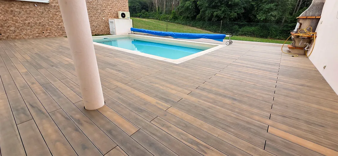 Terrasse composite Natural Beech continue depuis l'espace couvert jusqu'à la piscine et au barbecue, colonne blanche et pelouse en arrière-plan.