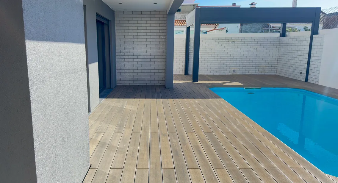 Terrasse composite CDECK® Natural Beech reliant l'espace couvert de la maison à la piscine, pergola anthracite contre le mur en briques blanches.