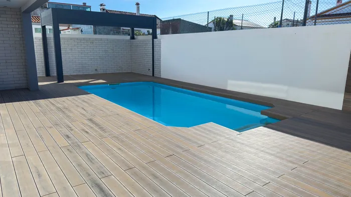 Patio con piscina rodeada de tarima composite CDECK® Natural Beech, pérgola antracita al fondo y muros blancos.