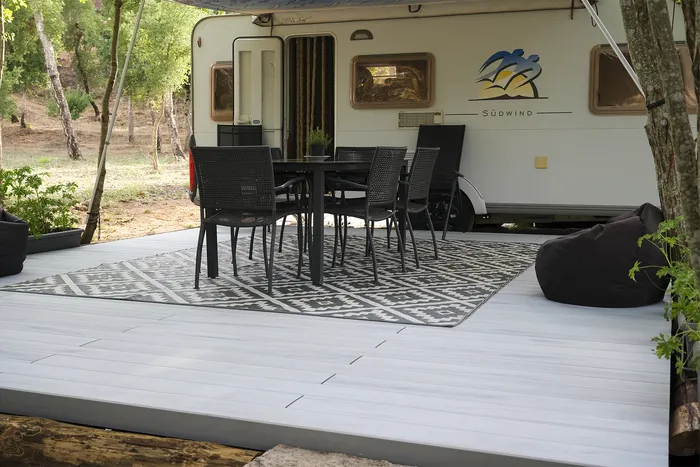 Salon d'extérieur dressé face à la caravane, tapis graphique et chaises noires sous les arbres.