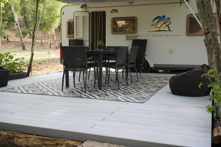 Salon d'extérieur dressé face à la caravane, tapis graphique et chaises noires sous les arbres.