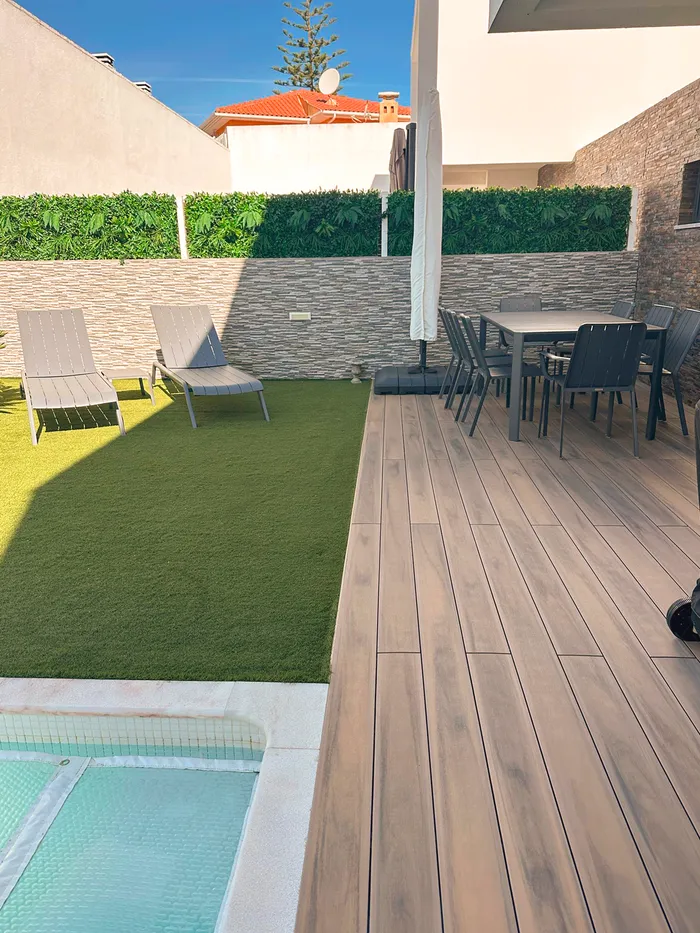 Deck compósito CDECK® Natural Beech junto à piscina, com mesa de refeições, relvado com espreguiçadeiras e muro de pedra.