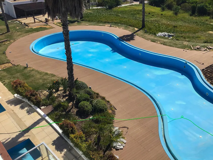 Piscine sinueuse turquoise cernée de terrasse composite et de palmiers dans un jardin.