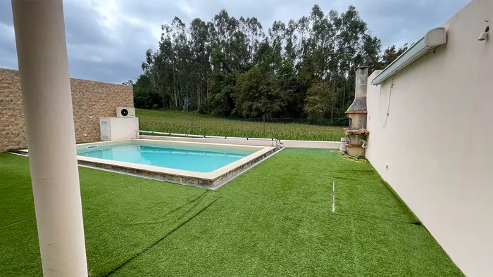 Piscine entourée de gazon synthétique avant les travaux, barbecue contre le mur blanc et eucalyptus en arrière-plan.