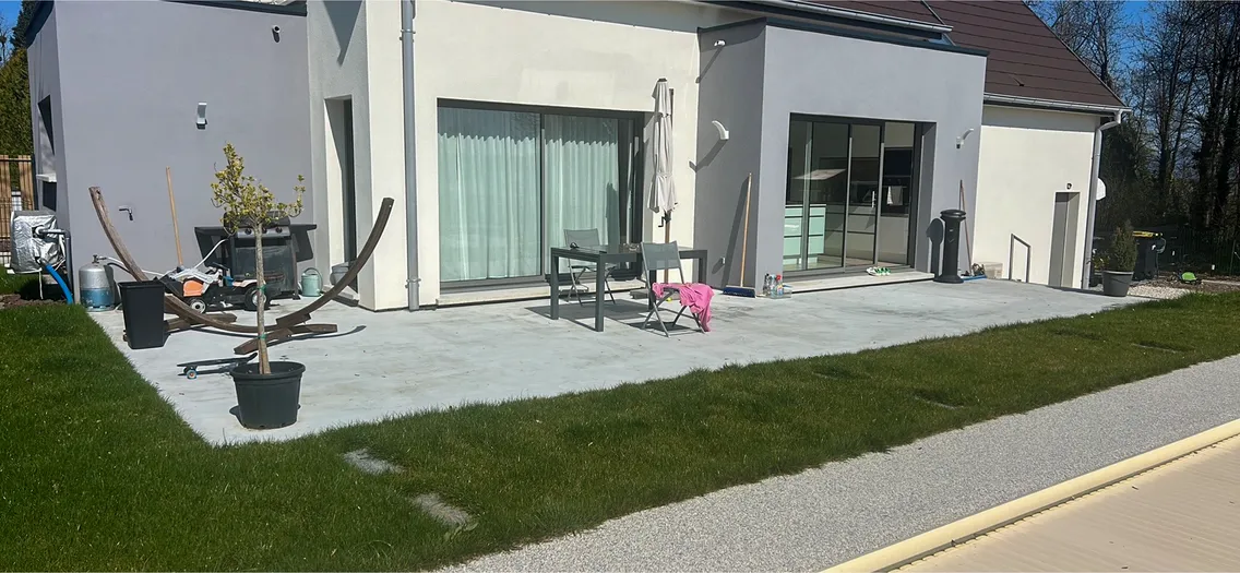 Terrasse en dalle béton avant les travaux, avec table, chaises, barbecue et pots contre la façade de la maison, pelouse au premier plan.