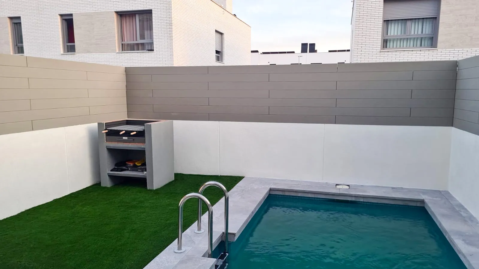 Garden with a swimming pool enclosed by walls clad in CWALL® Grey, with a barbecue and lawn, in Torrejón de Ardoz.